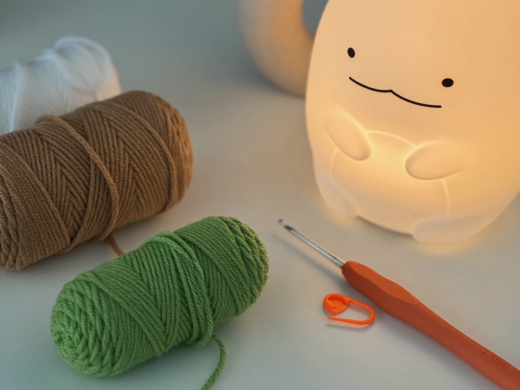 Crochet yarn, hook, and stitch marker on a table under a warm glowing lamp.